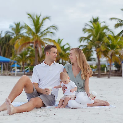 A family of three sits on a sandy beach with palm trees and blue canopies in the background, enjoying a relaxed moment together.